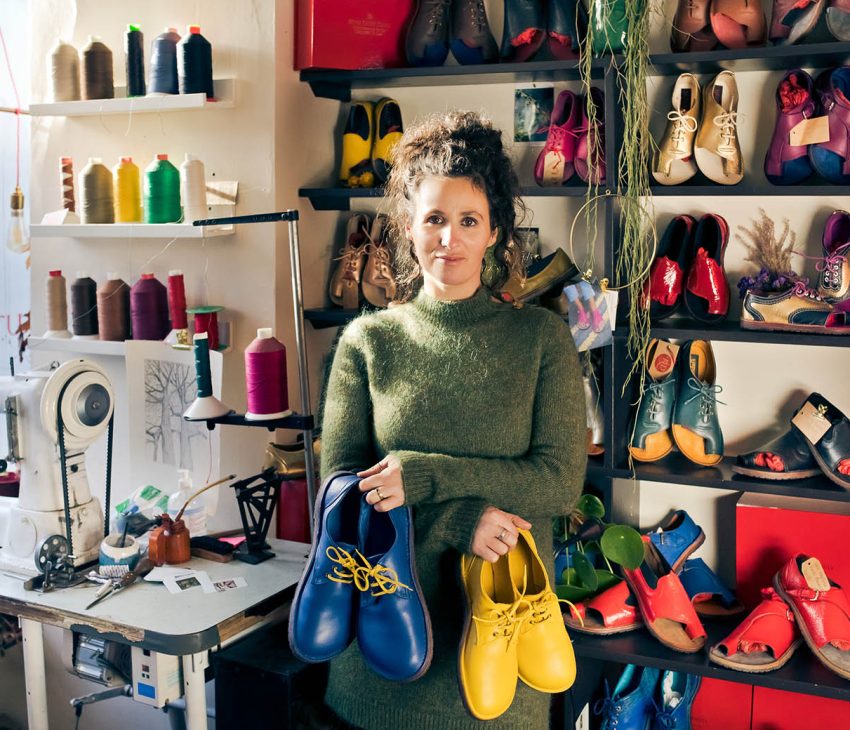 Woman in green jumper holding two pairs of shoes in a workshop with shelves of shoes behind her